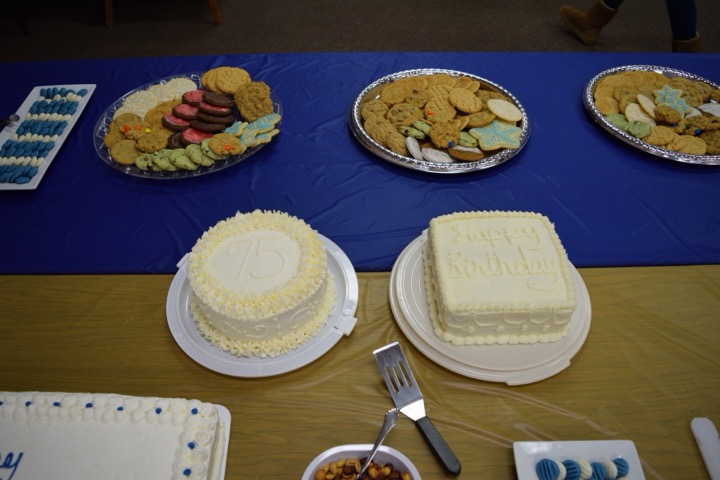 Two white birthday cakes marked 75 beside platters of cookies and treats